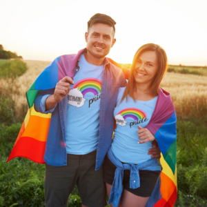 A man and a women holding a rainbow flag wearing blue Richmond Pride shirts that have the shape of Madison County Kentucky with a rainbow coming out of it.